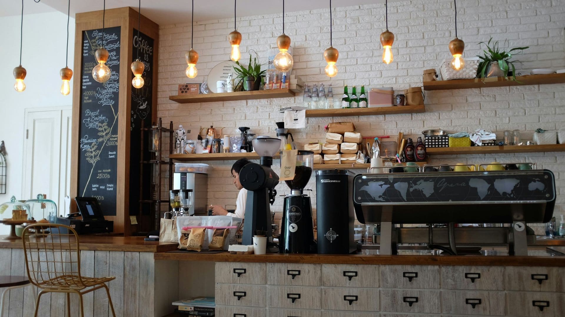 Barra de cafetería con máquina de espresso, muro de ladrillo pintado y lámparas colgantes