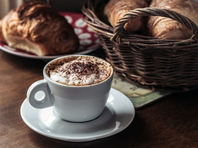 Taza de capuchino con cacao espolvoreado junto a un canasto de croissants
